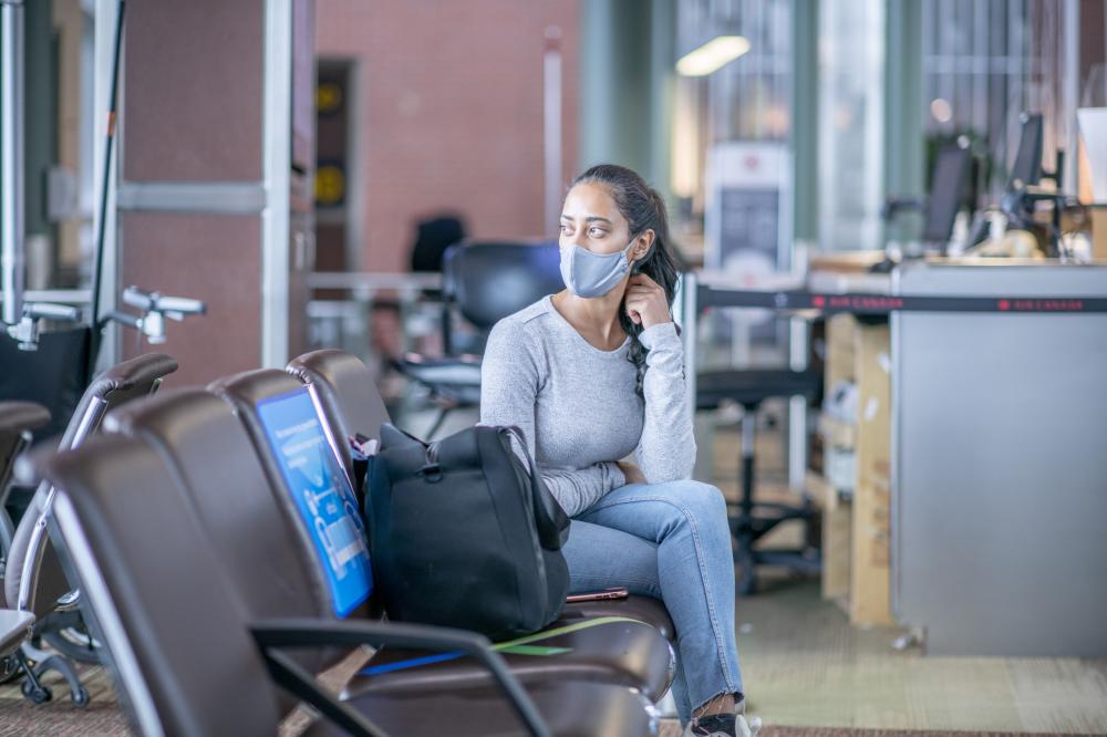 A young woman of Middle Eastern ethnicity is sitting at the airport departure gate. She is wearing a face mask to prevent the spread of germs during the coronavirus pandemic.